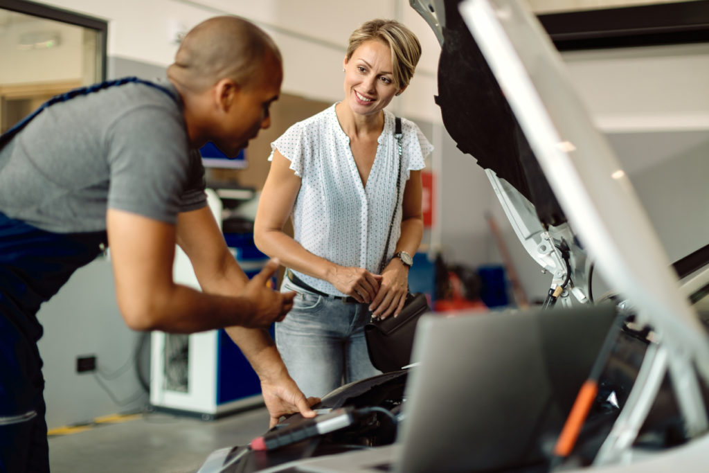 Young happy woman communicating with her auto repairman who is examining engine problems in a workshop.