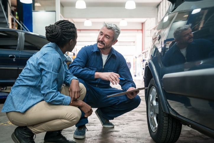 Car mechanic checking car wheel in the auto repair shop, holding clipboard and talking with customer.