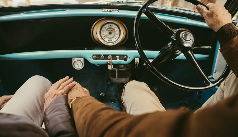 Araba sürerken el ele tutuşan kıdemli bir çiftin yakın çekimi.  Elini tutan yolcu koltuğunda oturan kadınla arabayı kullanan yaşlı adam.