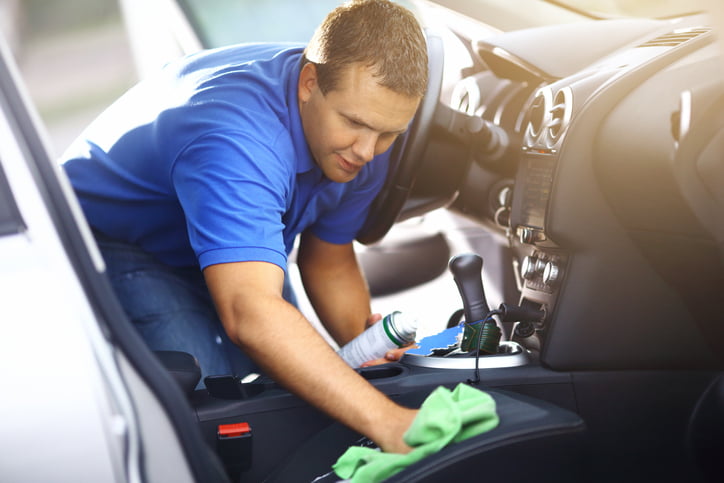 Adult caucasian man cleaning passenger seat with a green cloth and cleaning product. He applied some cleaning foam onto the seat. The guy has short brown hair and wearing blue polo shirt