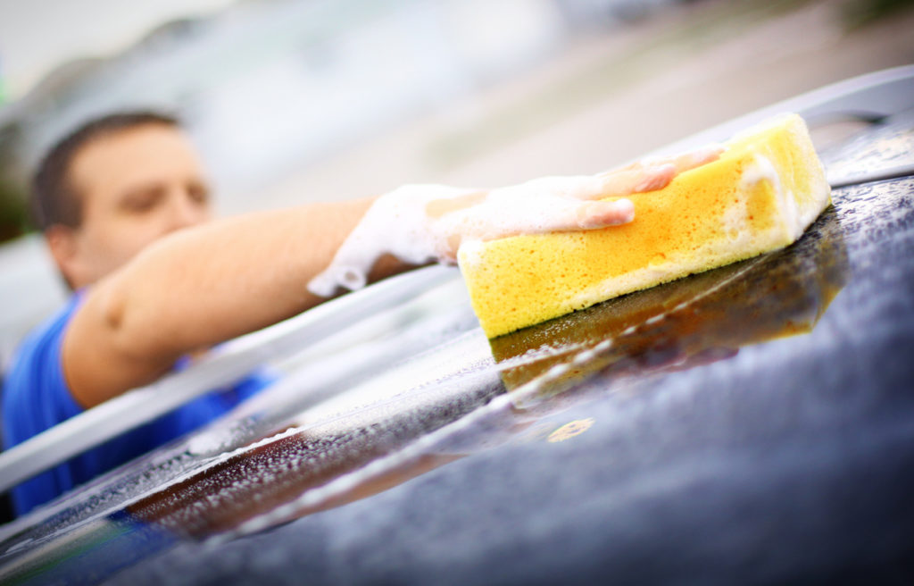 Unrecognizable middle aged man washing his car with yellow sponge and car wash shampoo.He's cleaning car roof.Hand and sponge in focus,he's blurry.He's wearing blue polo shirt.Copy space.