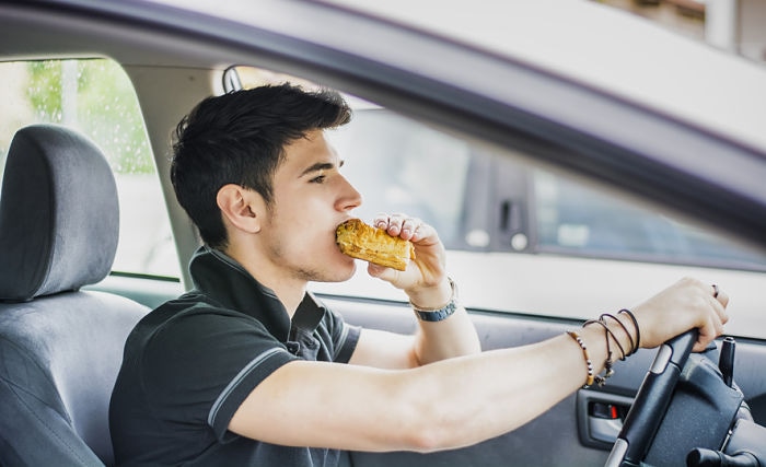 Young handsome man driving his car while eating food in the traffic